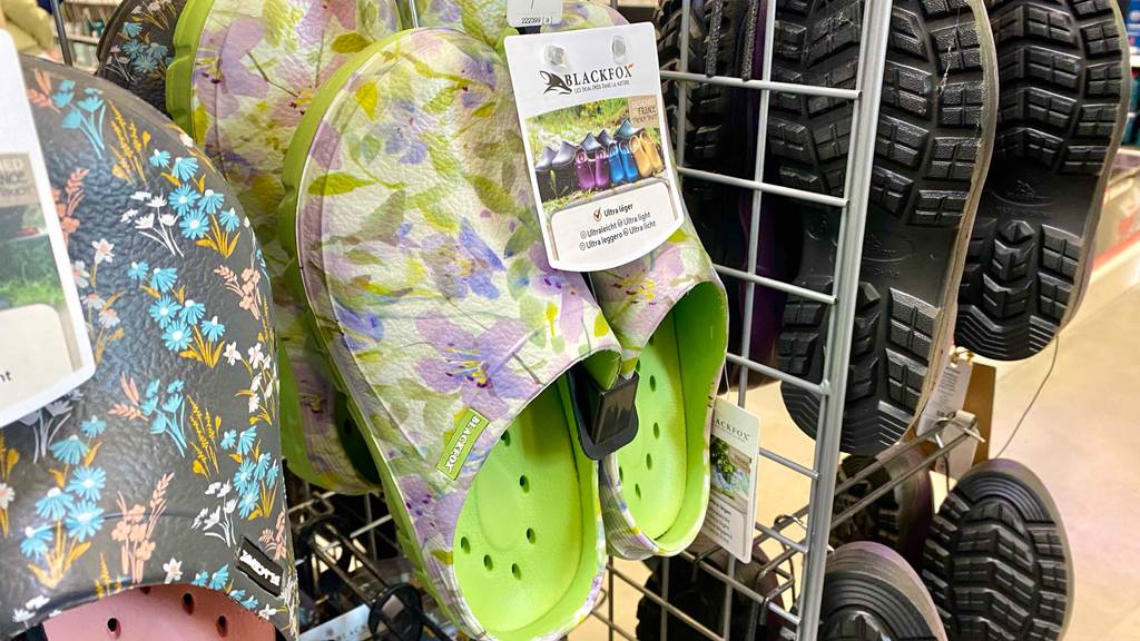 Blackfox Gartenschuhe im Test: Kunststoff-Gartenschlappen im dezenten floralen Design mit grüner, gelöcherter Innensohle hängen zwischen anderen Modellen an einem Verkaufsstand.