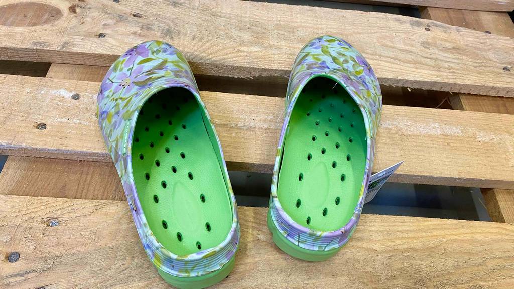 Gartenschuhe-Test: Ein Paar Kunststoff-Gartenschlappen in floralem Design mit grüner, gelöcherter Innensohle stehen auf Holzbrettern.