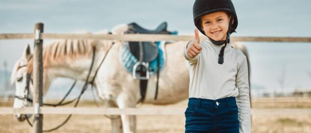 Reithose-Kinder-Test: Ein Mädchen in blauer Reithose und mit Helm steht im Vordergrund, hält den Daumen nach oben, trägt einen Reithelm. Dahinter ist ein gesatteltes Pferd zu sehen.