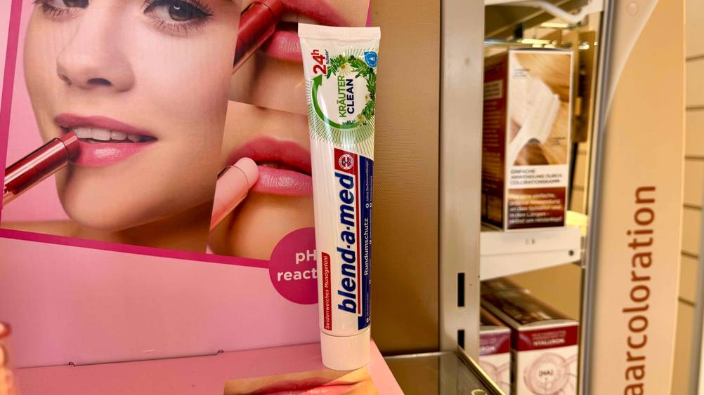 Vor einem Plakat steht eine Tube der getesteten Blend-a-med-Zahnpasta.