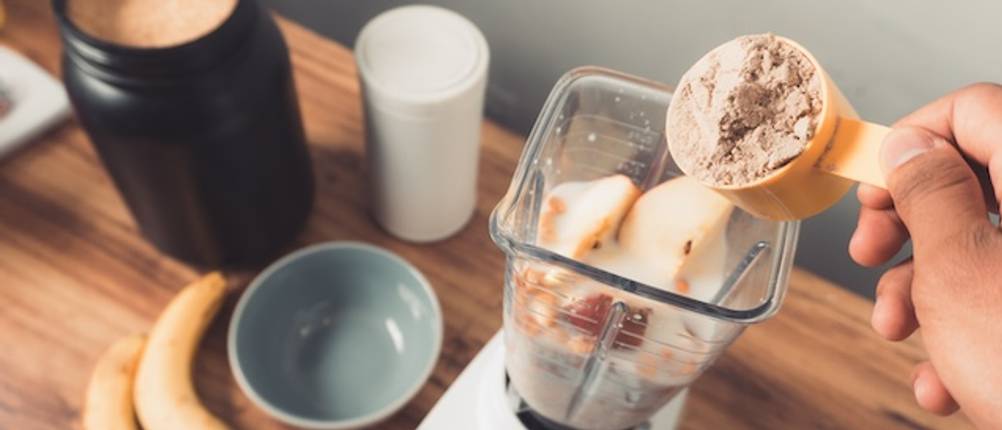Shakes im Mixer-Test: Eine Hand füllt Proteinpulver in einen eckigen Mixer-Behälter, in dem sich bereits Milch und Äpfel befinden. Der Mixer steht auf einer Arbeitsplatte aus Holz.