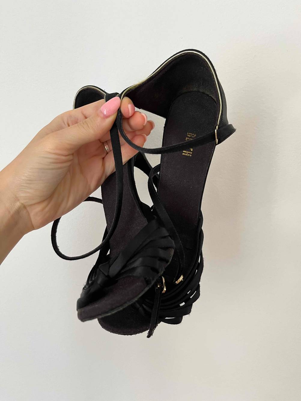 Eine Hand hält ein Paar getestete Tanzschuhe an den Riemchen vor einer weißen Wand.
