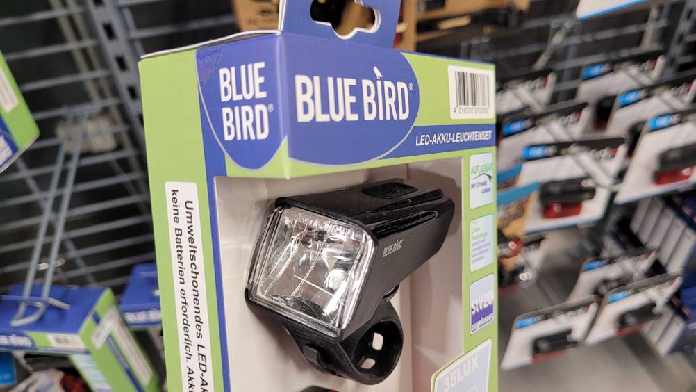 Im Test. Fahrradlampen-Set der Marke Blue Bird, mit Fokus auf die Vorderlampe in einer hellgrün-blauen Verpackung hängt in einem Verkaufsbereich.