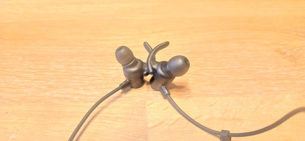 Nahaufnahme der getesteten Bluetooth-Sportkopfhörer inklusive der Ohrenbügel auf einer Holzfläche.
