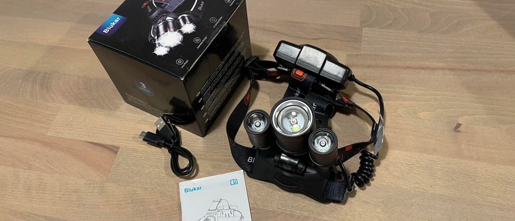 Die Stirnlampe Blukar K9116 liegt ausgepackt neben Verpackung, USB-Kabel und Anleitung auf einem Holztisch. Zu sehen sind das Leuchtmodul mit drei LEDs sowie das Kopfband mit Akkupack.
