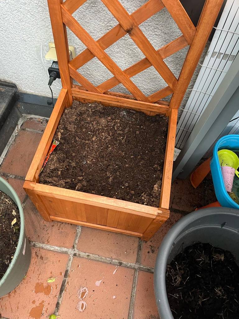 Ein aus Holz gefertigter Blumenkasten mit Rankgitter im Test steht auf einem Balkon neben weiteren Blumentöpfen.