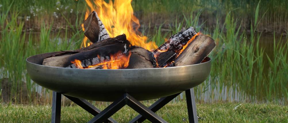 Blumfeldt-Feuerschale-Test: Eine Feuerschale mit brennendem Scheitholz auf einer Grünfläche vor einem kleinen See.