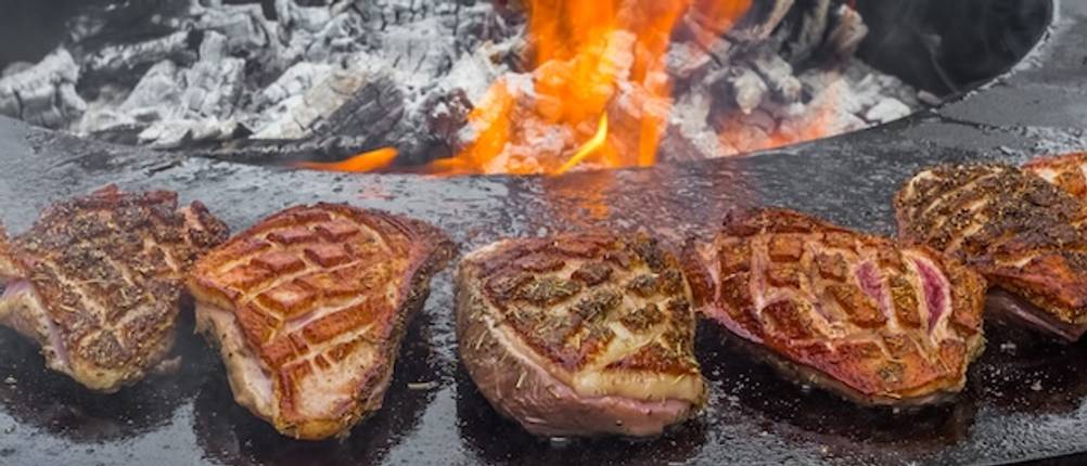 Blumfeldt-Feuerschale getestet: Fertig gegrillte Steaks liegen auf dem Rand einer runden, schwarzen Feuerschale mit brennender Glut.