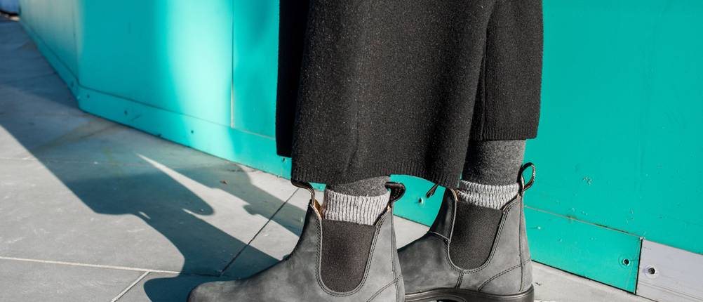 Blundstone-Test: Frau mit langen schwarzem Rock trägt Boots mit Einstiegschlaufen vor grüner Wand.