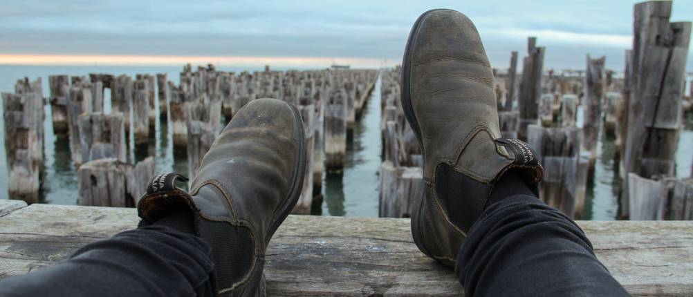 Blundstones im Test: Mann legt seine Beine hoch auf einem Holzbalken, mit Blick aufs Meer.