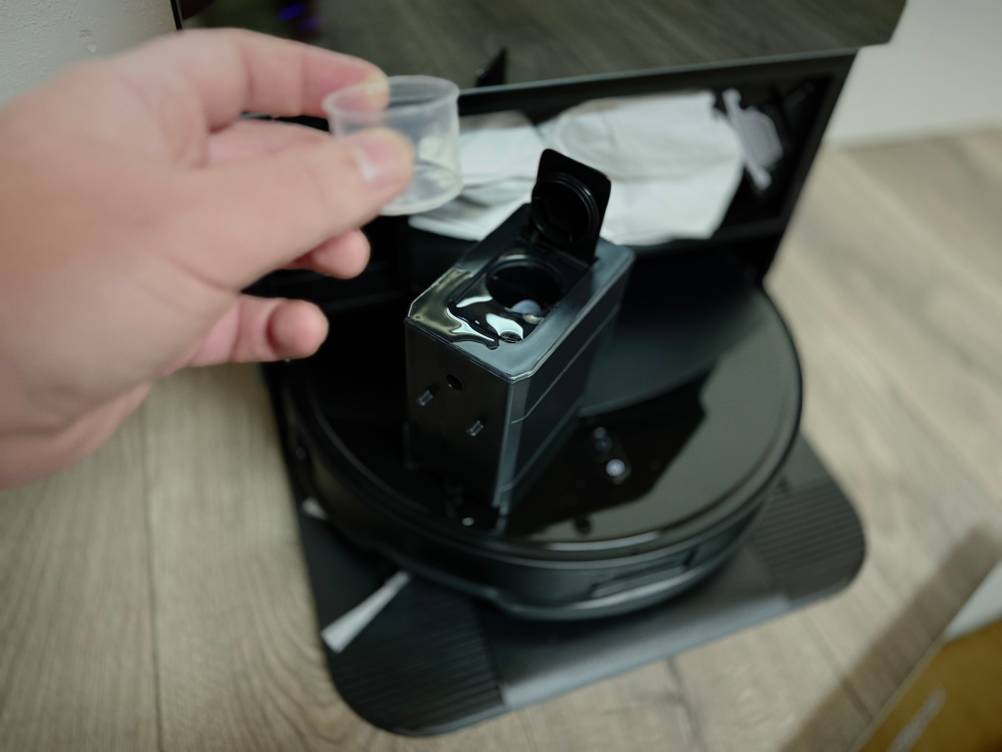 Eine Hand hält eine Dosierkappe vor einem Saug-Wisch-Roboter.