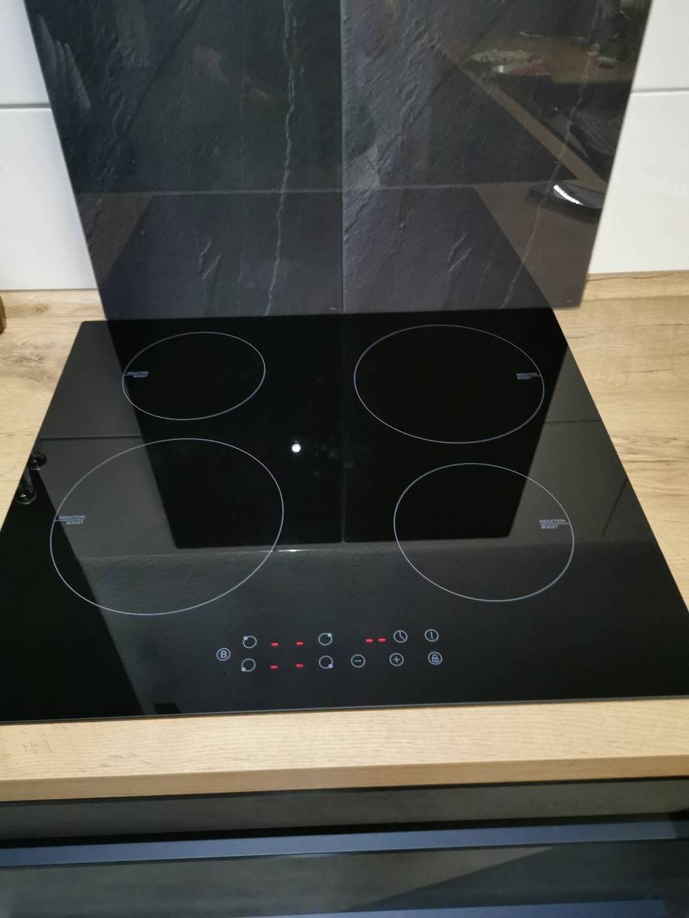 Ein schwarzes Induktionskochfeld im Test, inklusive vier Kochzonen auf einer Küchenarbeitsplatte aus hellem Holz, sowie ein rückwärtiger Spritzplatte auf weißen Fliesen.