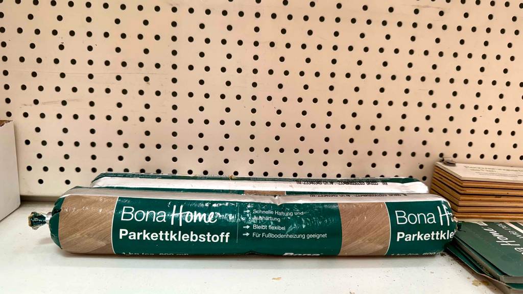 Im Test: Ein verpackter Parkettkleber liegt in vor einer Lochwand.