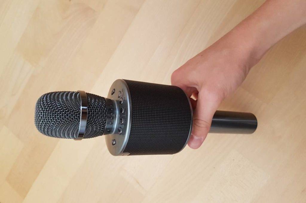 Karaoke-Anlage-Test: Nahaufnahme einer Hand vor hellem Holzboden, die ein schwarz-silbernes Mikrofon mit integriertem Lautsprecher festhält.