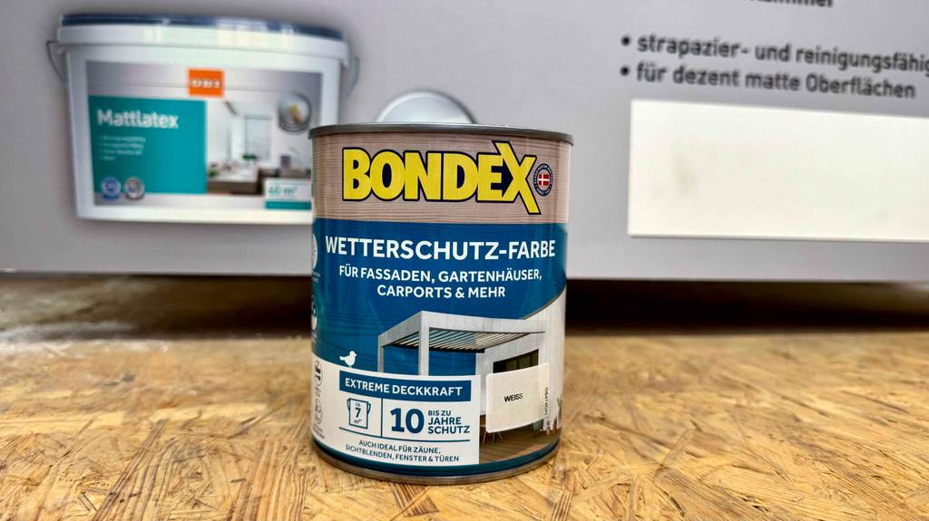 Die getestete weiße Wetterschutzfarbe in der Dose steht vor einem Plakat mit Farbprodukten.
