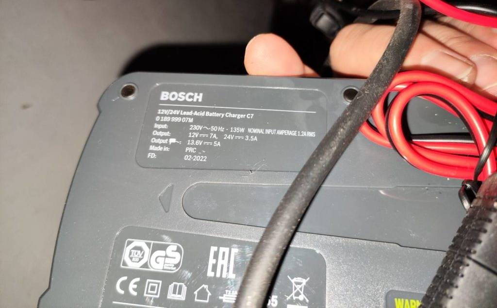 Nahaufnahme einer Hand an der Rückseite inklusive Typenschild von einem getesteten Autobatterie-Ladegerät der Marke Bosch unter rotem und schwarzem Kabel.