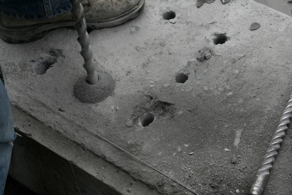 Bosch-Akku-Bohrhammer im Test: Ein Fuß steht auf einer Betonplatte und ein großer Bohrer bohrt das sechste Loch in diese Platte. Ein weiterer Bohrer liegt daneben.