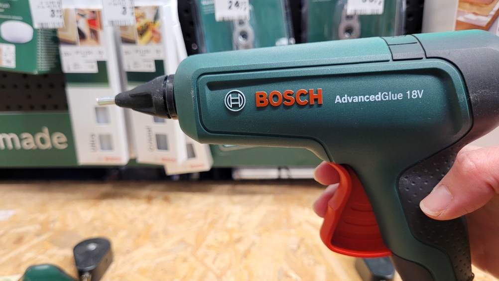 Heißklebepistole mit Akku wird in einem Geschäft in der Hand gehalten, Verpackungen im Hintergrund sichtbar