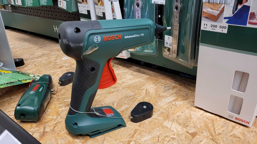 Heißklebepistole mit Akku steht auf einer Holzplatte in einem Verkaufsregal neben Zubehörteilen.