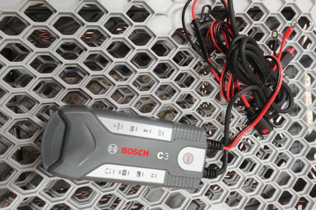 Bosch-C3-Autobatterie-Ladegerät im Test liegt inklusive Kabel auf einem gelöcherten, hellgrauen Untergrund.