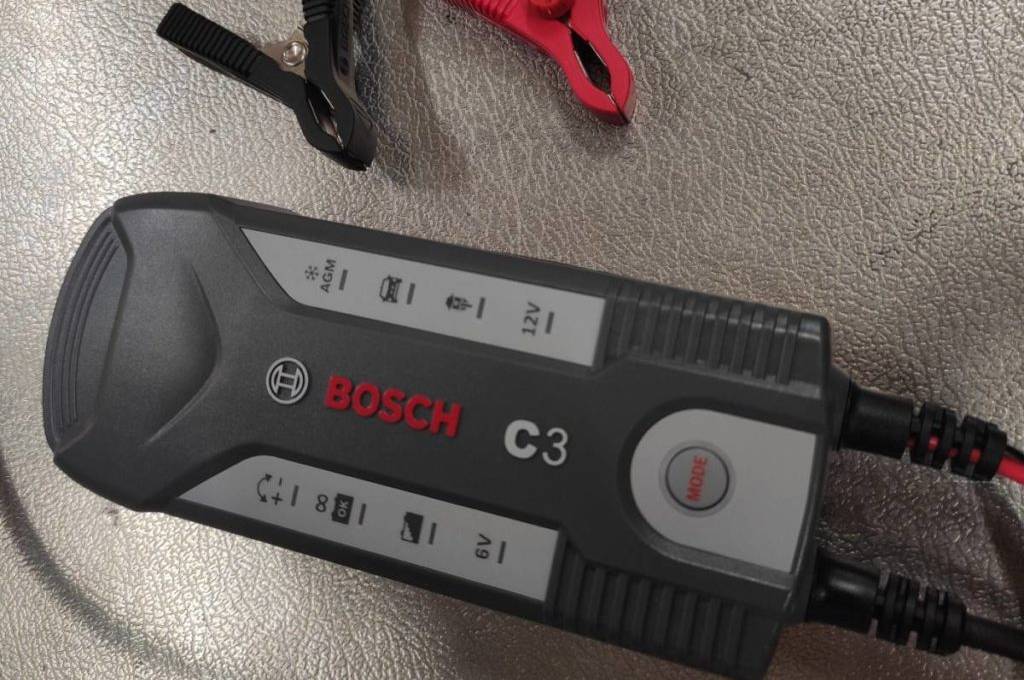 Getestetes Autobatterie-Ladegerät C3 von Bosch liegt neben roter und schwarzer Pol-Klemme auf einem grauen Untergrund.