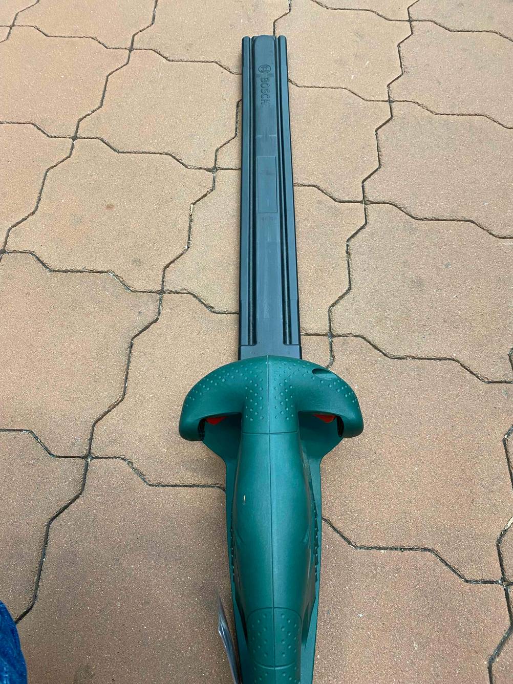 Im Test: Eine Bosch Heckenschere mit einem Schutzüberzug über der Schneide auf einem Pflasterboden von oben.