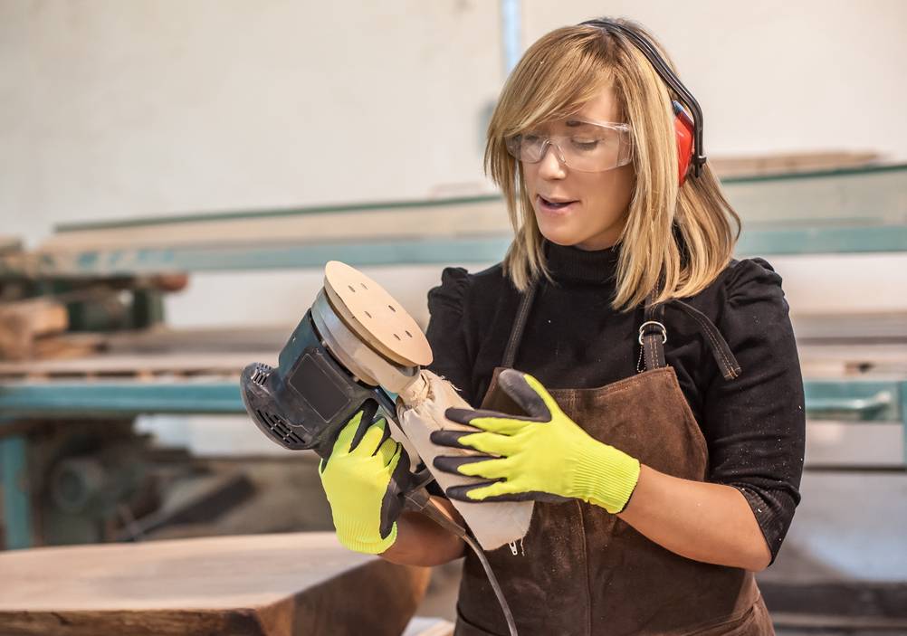 Bosch-Exzenterschleifer im Test: Eine junge Frau mit Schutzbrille und Gehörschutz schaut von unten auf den Schleifteller.