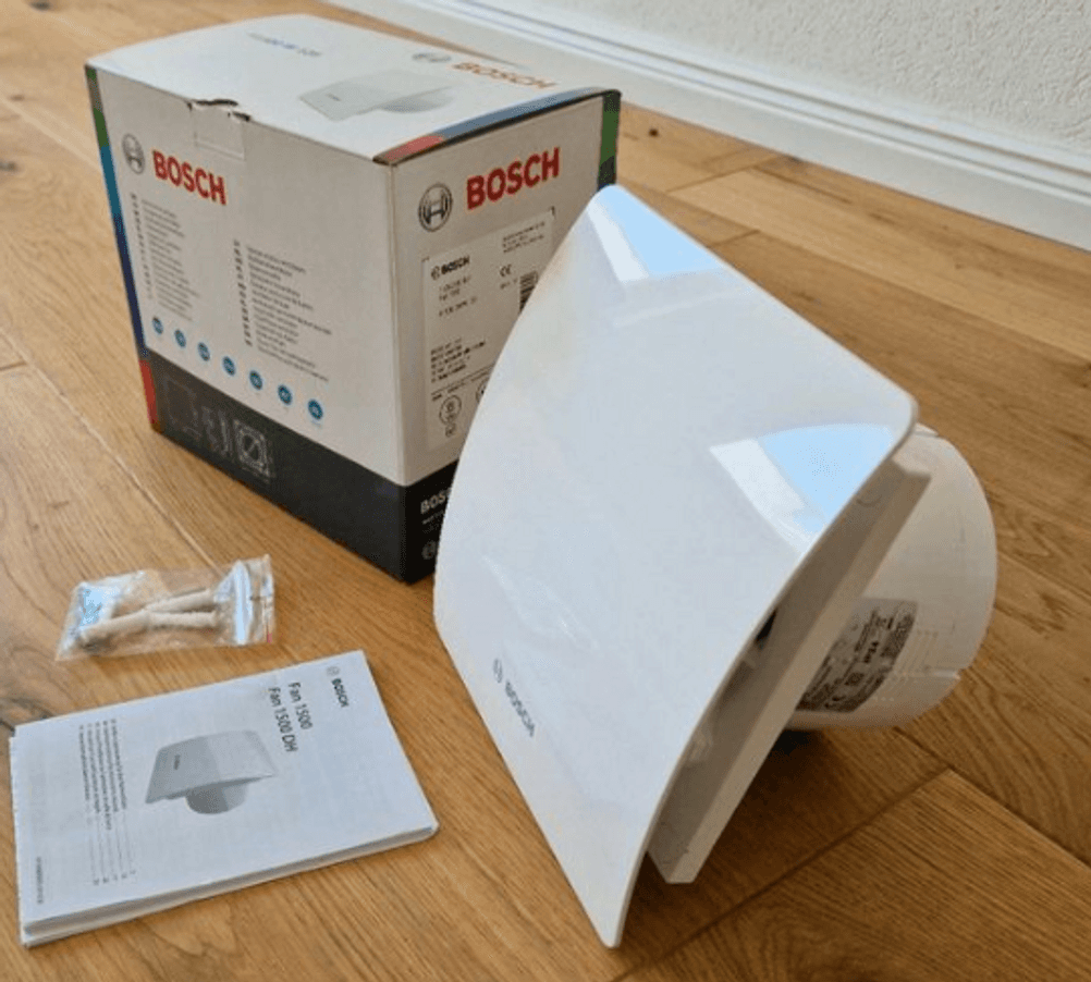 Ansicht von schräg oben auf einen getesteten Bosch Badlüfter, der sich neben seiner Originalverpackung, dem Montagematerial und der Betriebsanleitung auf einem Holzboden befindet.