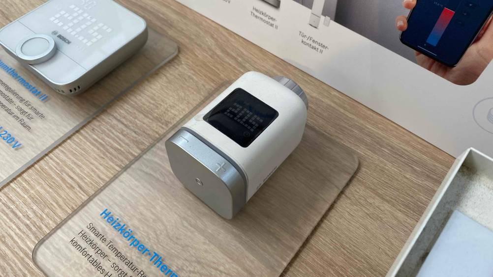 Heizkörperthermostat-Test: Ein Gerät von schräg seitlich auf einer transparenten Infotafel im Hintergrund eine anderes Gerät.