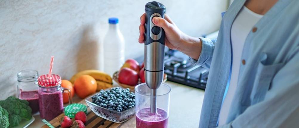 Bosch-Mixer im Smoothie-Test: Eine Damenhand bedient einen Stabmixer auf Knopfdruck und mixt ein violettfarbenes Getränk. Im Hintergrund sind Blaubeeren und weitere Früchte zu sehen.