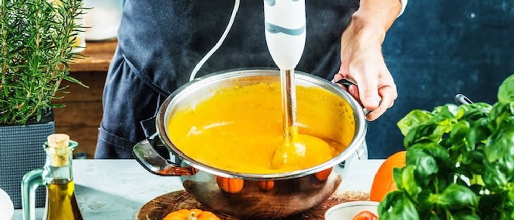 Das Zubehör getestet: Mit einem Stabmixer wird eine gelbe Suppe in einer Edelstahlschüssel zubereitet.