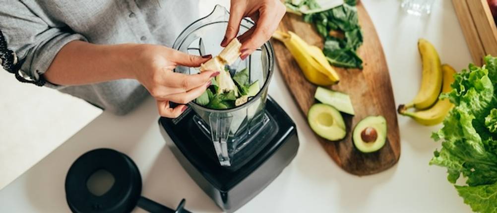 Bosch-Mixer getestet: Eine Frau füllt Bananen in einen Mixbehälter, in dem sich bereits grüne Zutaten befinden. Auf einem Holzbrett daneben liegen die Bananenschale und Avocadohälften.