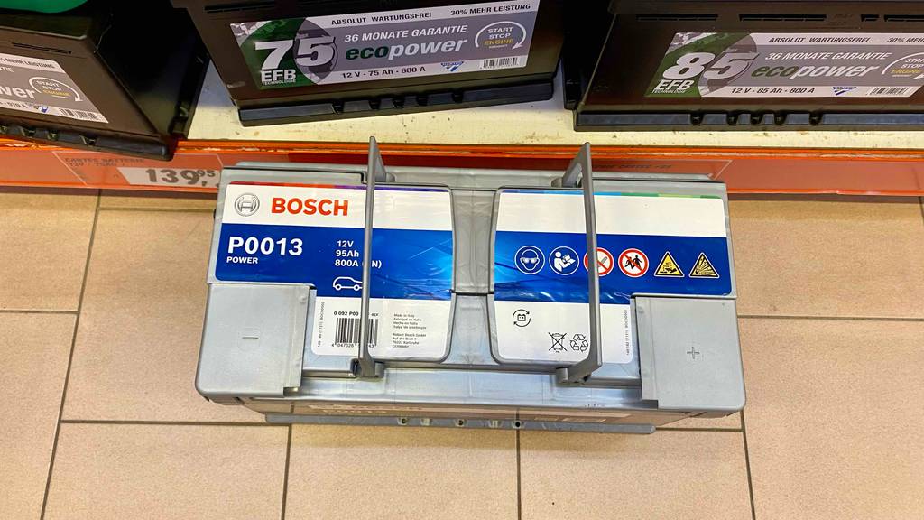 95-Ah-Autobatterien-Test: Ansicht von oben auf eine hellgraue Bosch-12-Volt-Autobatterie P0013 inklusive zweier Handgriffe vor einem Verkaufsregal mit anderen Autobatterien.