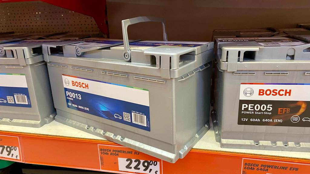 Im Test: eine 95-Ah-Autobatterie in Hellgrau der Marke Bosch, mit Fokus auf die beiden Tragegriffe steht zwischen anderen 12-Volt-Autobatterien auf einem Verkaufsregal.