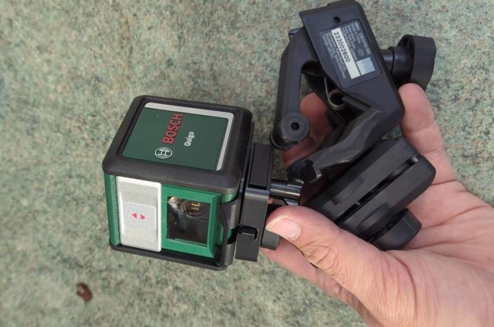 Batteriebetriebener, getesteter Kreuzlinienlaser von Bosch inklusive Halterung in einer linken Hand vor grünlichem Hintergrund.