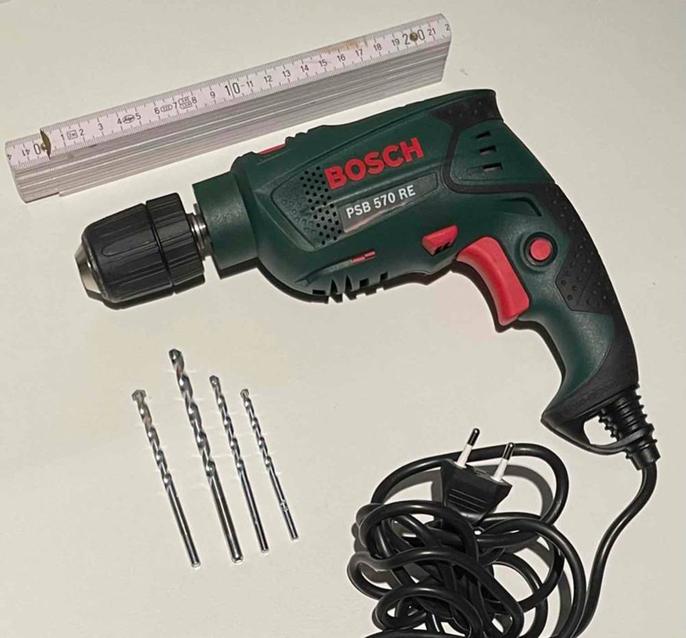 Elektrische PSB 570 RE Bosch-Schlagbohrmaschine im Test liegt inklusive vier Bohrspitzen und Zollstab auf einem hellen Untergrund.