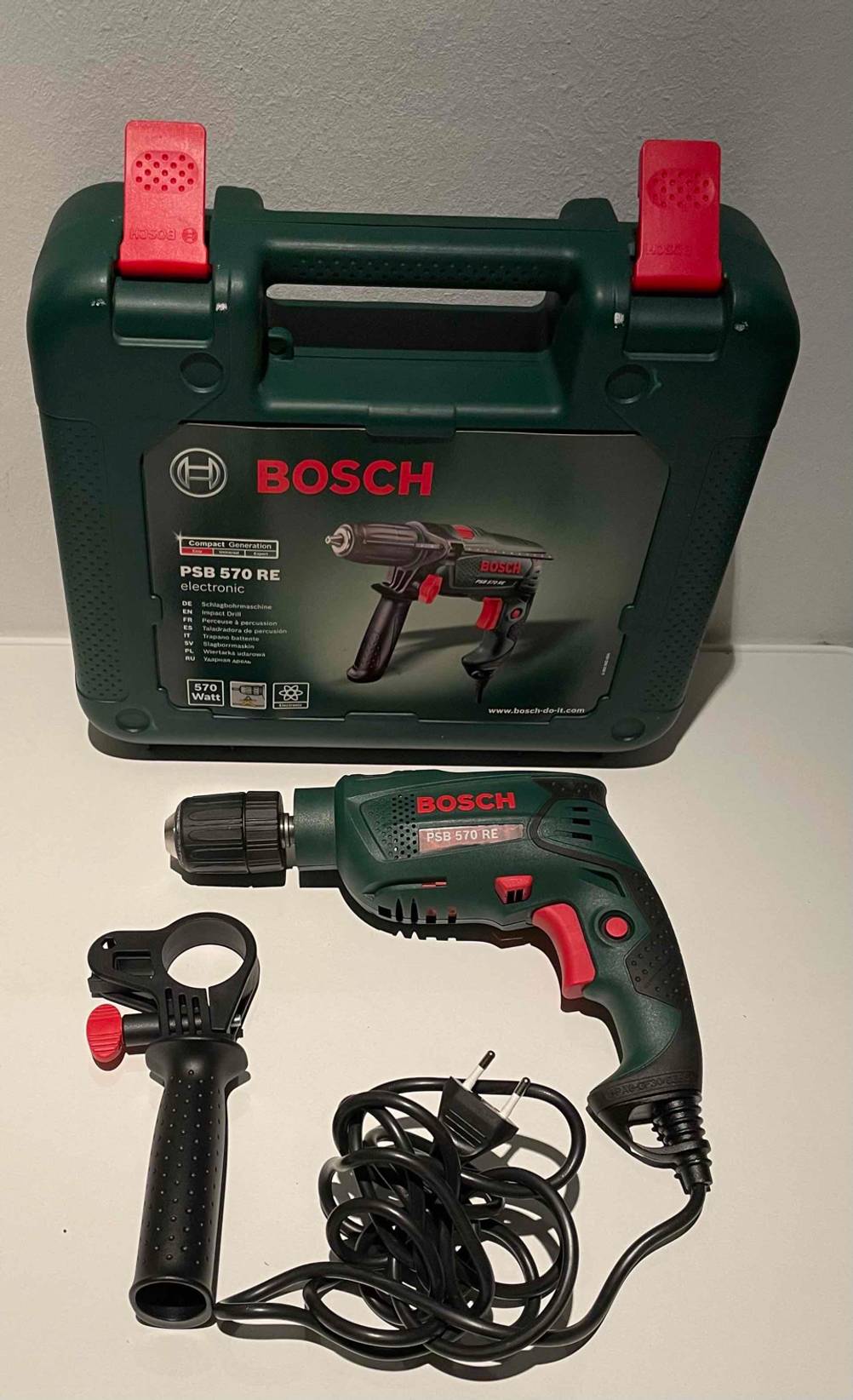 Elektrische, getestete Bosch-Schlagbohrmaschine befindet sich vor ihrem Transportkoffer inklusive Zusatzhandgriff auf einer hellen Fläche.