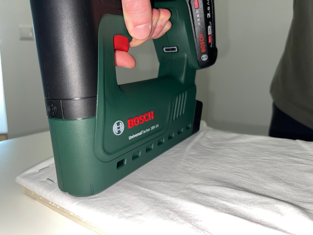 Der Bosch UniversalTacker 18V‑14 tackert weißen Stoff auf eine Holzplatte. Die Auslösung erfolgt manuell über den roten Knopf.