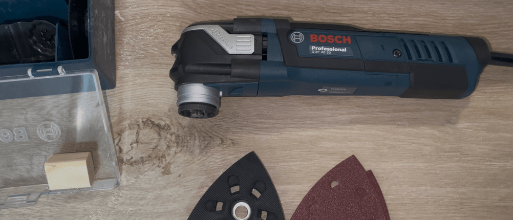 Das Bosch GOP 40-30 liegt auf einer Holzplatte, daneben sind Schleifaufsatz und Koffer sichtbar. Alle Teile sind sauber angeordnet.