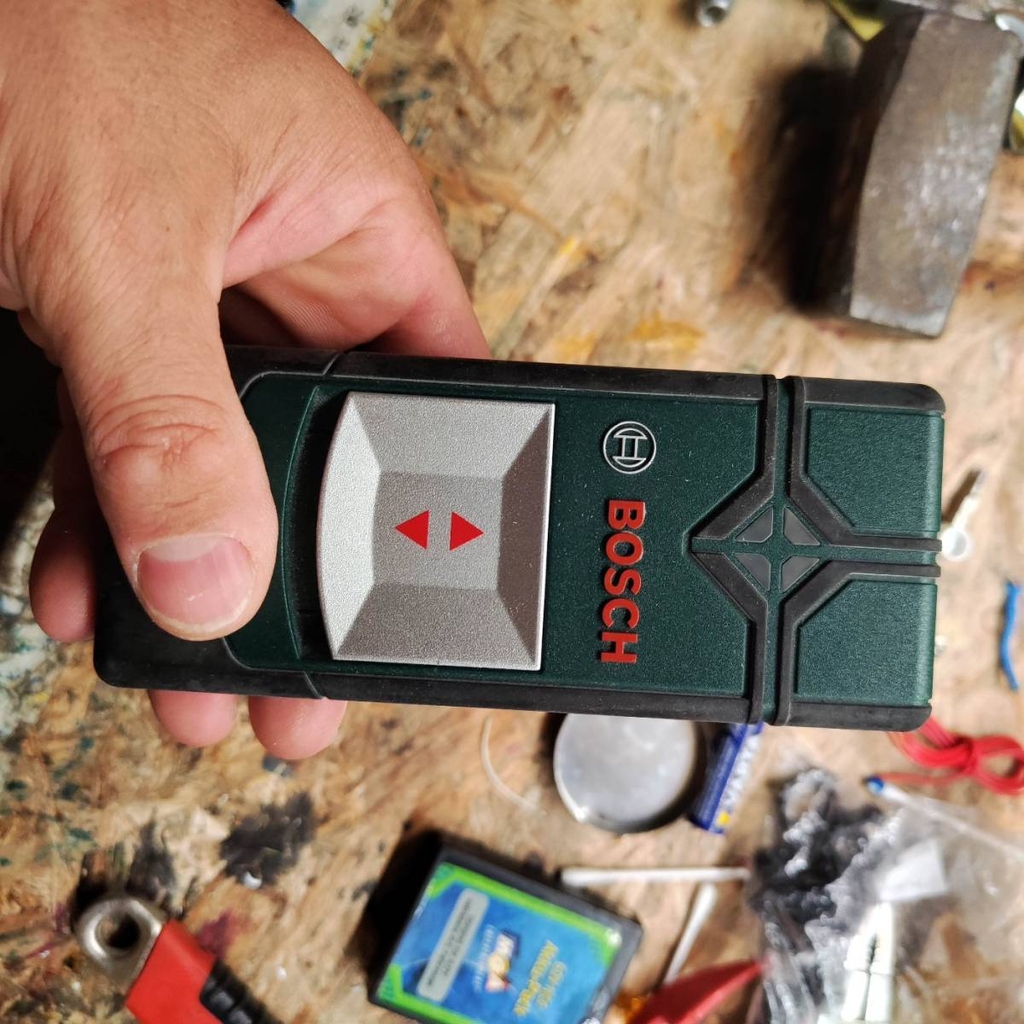 Hand hält einen getesteten Leitungssucher der Marke Bosch vor Hintergrund mit Werkzeugen.
