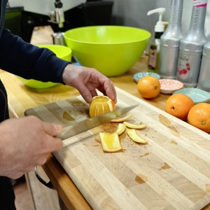 Eine Person schält eine Orange mit einem Messer auf einem Holzbrett, um sie für den Bosch VitaJuice 3 MES3500 vorzubereiten. Weitere Orangen und grüne Schüsseln stehen im Hintergrund.