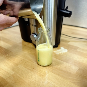 Frisch gepresster Saft wird aus einem Edelstahl-Messbecher in eine Glasflasche gegossen. Der Bosch VitaJuice 3 MES3500 steht im Hintergrund auf einer Küchenarbeitsplatte.