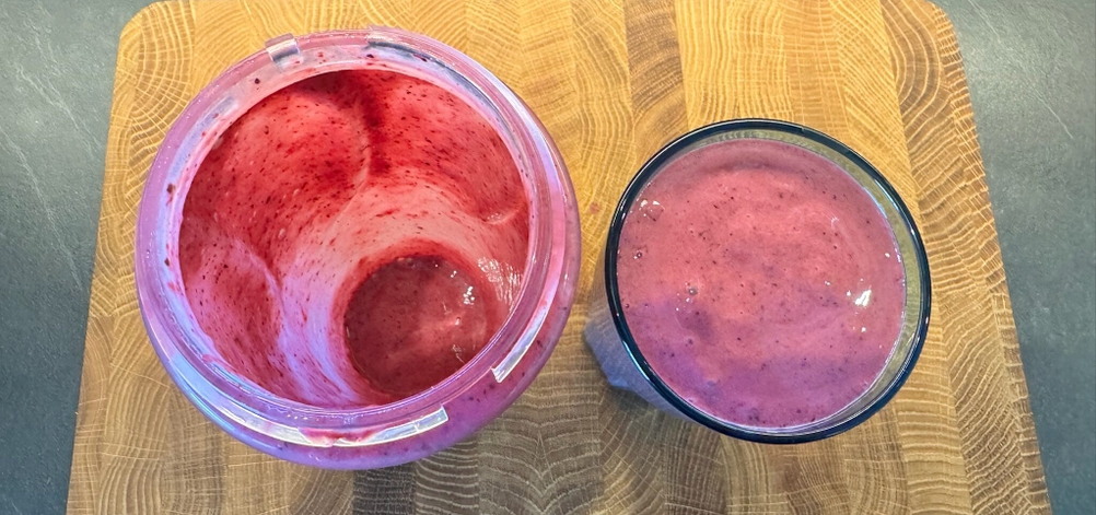 Neben dem leeren, benutzten Behälter des Smoothie-Makers steht ein gefülltes Smoothie-Glas.