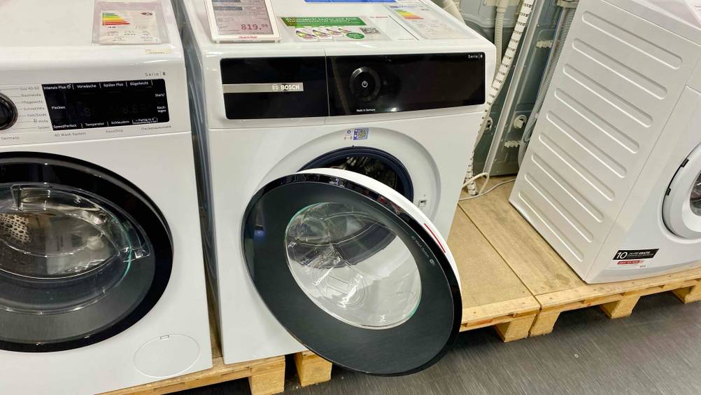 Waschmaschine 10 kg getestet: Ein Gerät mit geöffneter Klappe auf eine Holzsockel neben anderen Geräten in einem Verkaufsraum.