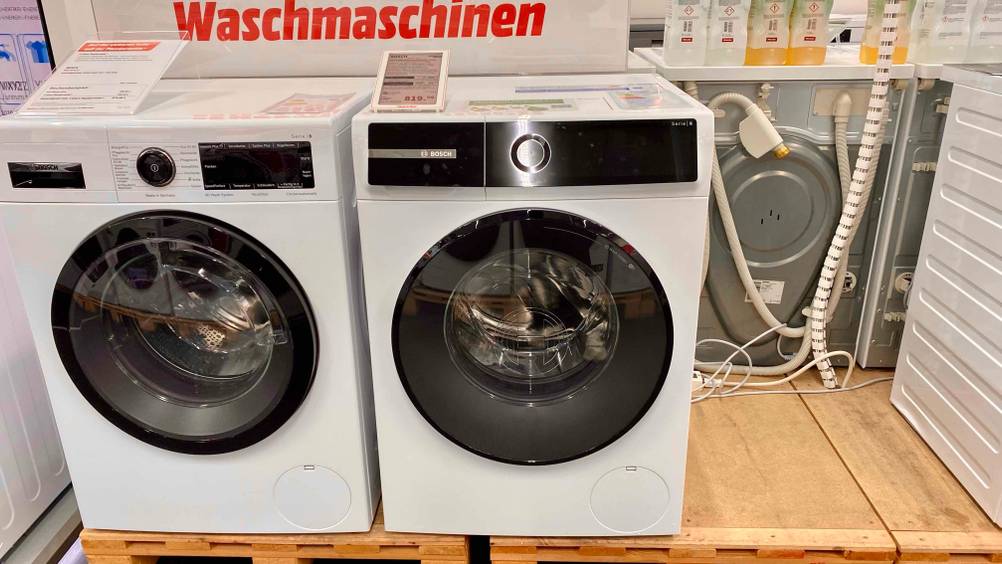 Eine getestete Waschmaschine 10 kg auf einem Holzsockel neben einem anderen Gerät in einer Verkaufsfläche, darauf steht ein Preisschild.