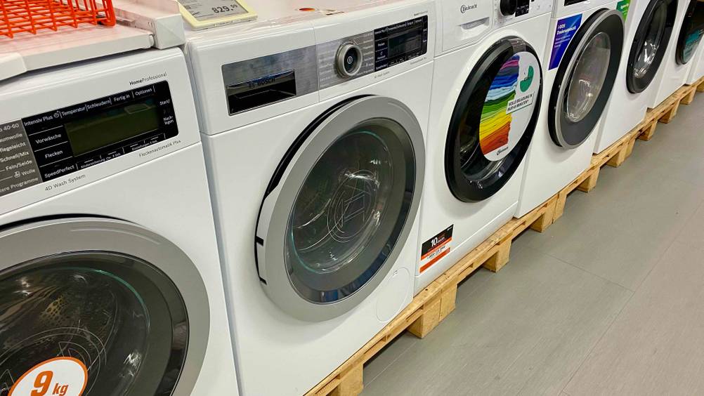 In einer Ausstellungsfläche steht eine getestete Bosch-Waschmaschine zwischen anderen Geräten auf Holzpaletten mit Preisangaben.