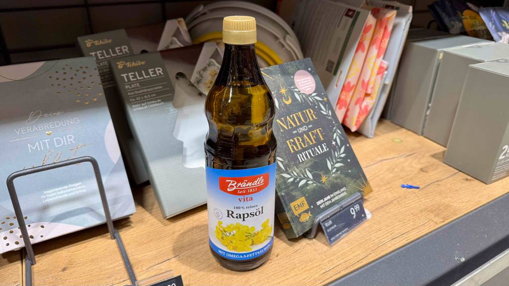 Rapsöl im Test: Eine Flasche steht vor verpackten Tellern in einem Holzregal.