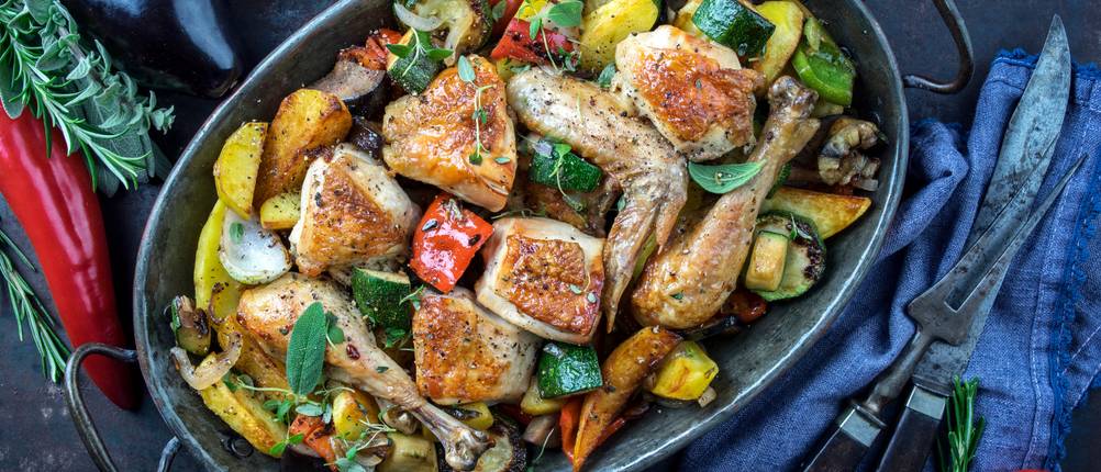 Ovale Bräter im Test: mit Gemüse, Kräutern und Fleisch befüllter ovaler Bräter auf einer dunklen Arbeitsplatte, verschiedene Utensilien, Tomaten und Rosmarin daneben platziert 