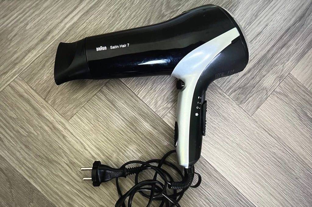 Nahaufnahme eines Ionen-Haartrockners im Schwarz-Silber-Design inklusive Kabel auf dem Boden mit Holzmaserung.