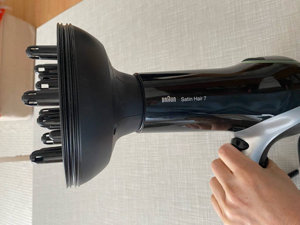 Profi-Haartrockner im Test: Hand hält Braun Satin Hair 7 mit einem schwarzen Diffusor über eine hellgraue Fläche.